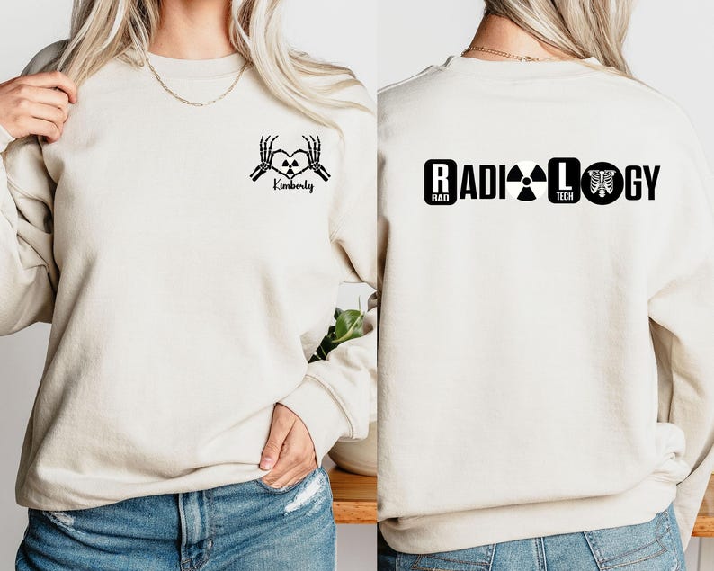 May include: Two cream-colored sweatshirts with black text and graphics. One sweatshirt has a skeleton hand heart graphic with the text "Kimberly". The other sweatshirt has the text "RADIOLOGY" with a radiation symbol, the text "RAD TECH", and a ribcage graphic.