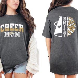 Pu&ograve; includere: Una maglietta grigia con la scritta "Cheer Mom" sul davanti e un megafono bianco con la scritta "Emily" sul retro. Il megafono &egrave; diviso a met&agrave; con un pompon dorato e bianco sul lato destro.