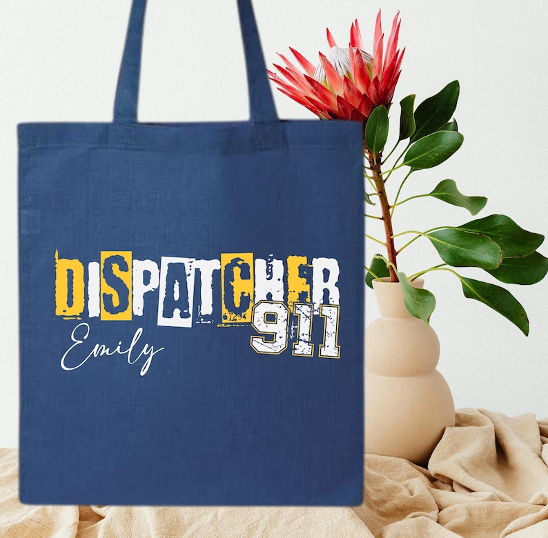 May include: A blue canvas tote bag with the text "Dispatcher 911" in yellow and white letters. The name "Emily" is written in white below the text.