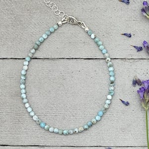 May include: A delicate beaded bracelet featuring small, round, light blue and white stones. The bracelet has a silver-colored clasp and is displayed on a light gray wooden surface, with sprigs of purple lavender.