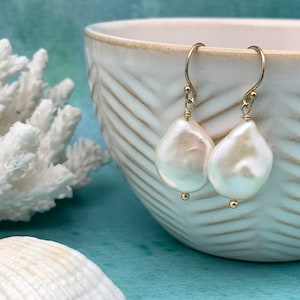 May include: A pair of gold-toned drop earrings with large, irregular white pearls. The earrings hang from delicate gold hooks. The background includes a white ceramic bowl, white coral, and a seashell, all set against a teal surface.