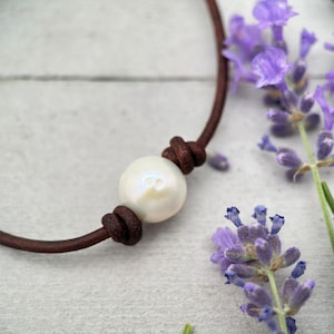 May include: A close-up of a pearl necklace with a brown leather cord. The pearl is white and iridescent. The necklace is knotted on both sides of the pearl. Lavender flowers are in the background.
