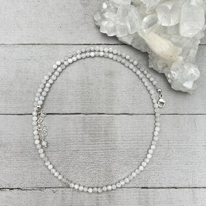 Puede incluir: Un collar de cuentas de piedra lunar blanca con un cierre plateado. El collar est&aacute; colocado sobre una superficie de madera blanca con un grupo de cristales blancos en el fondo.