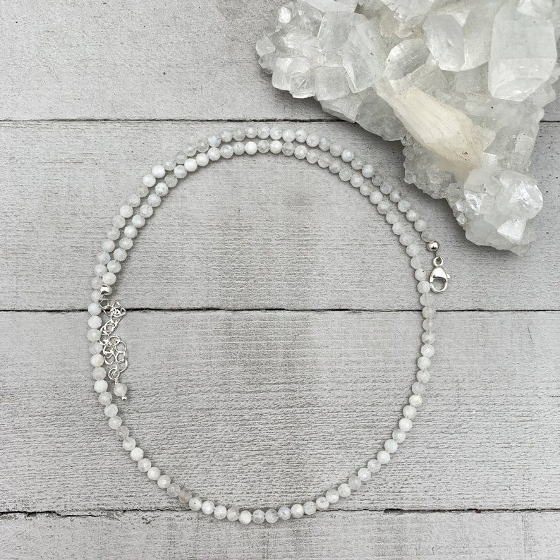 Puede incluir: Un collar de cuentas de piedra lunar blanca con un cierre plateado. El collar est&aacute; colocado sobre una superficie de madera blanca con un grupo de cristales blancos en el fondo.