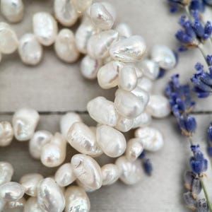 May include: A strand of white baroque pearls, with an irregular shape, arranged in a circular pattern on a light wood surface.
