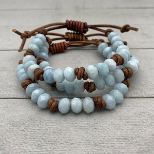 May include: A triple-wrap bracelet with light blue gemstone beads and brown leather cord. The bracelet has knots on the leather cord.
