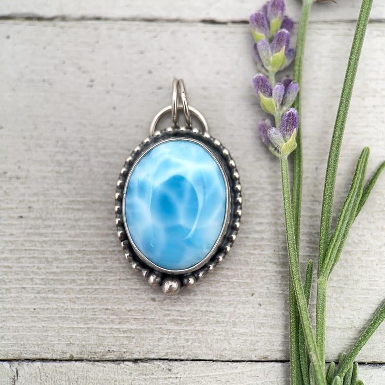 Puede incluir: Un colgante de forma ovalada con una piedra Larimar azul cielo pulida y patrones veteados en blanco. La piedra est&aacute; engastada en un marco de metal plateado con un borde de cuentas y una anilla circular en la parte superior para colgar.