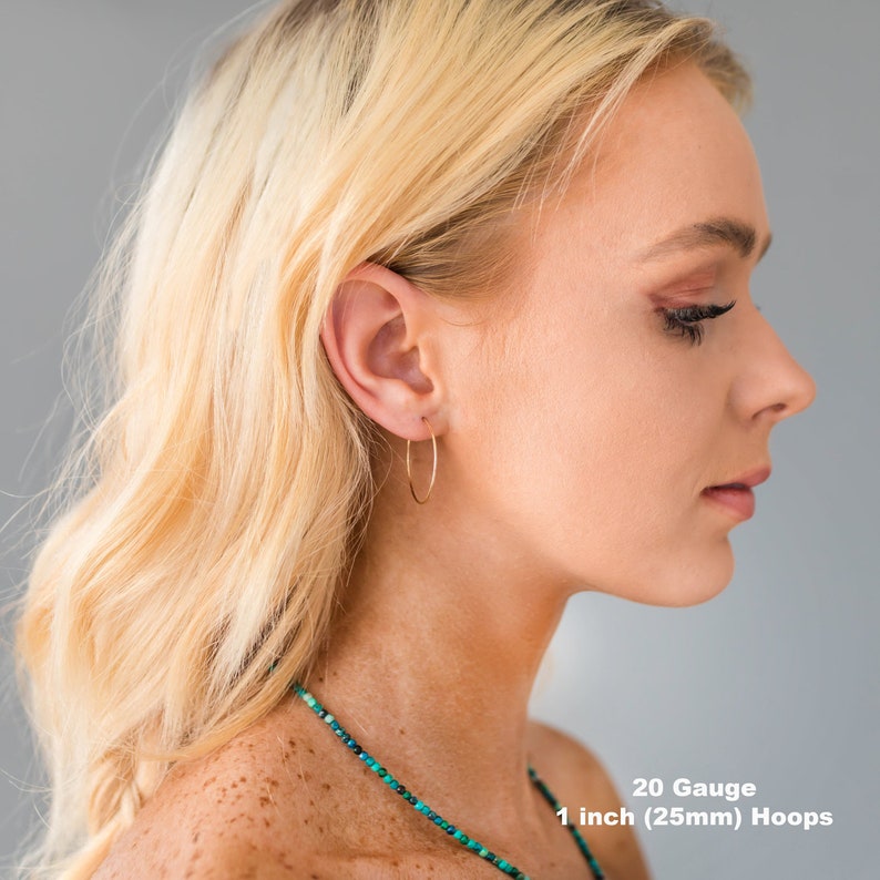 May include: A woman wearing gold-colored, thin hoop earrings. The earrings are 1 inch (25mm) in diameter and 20 gauge. The woman has blonde hair and is wearing a necklace.
