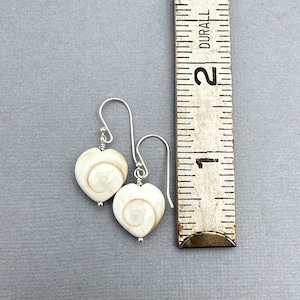May include: A pair of heart-shaped earrings with a spiral design in the center. The earrings are white with a light brown spiral and are suspended from silver-colored hooks. A ruler is next to the earrings.