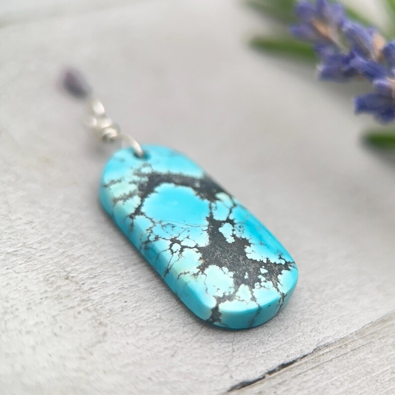 May include: A turquoise pendant with a silver bail. The pendant is a rectangular shape with a rounded top and bottom. The turquoise has a striking black matrix pattern. The background is a light grey surface.