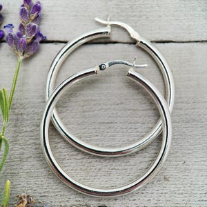 May include: A pair of polished silver hoop earrings with a smooth, rounded profile and a hinged post closure mechanism. The earrings are circular in shape and feature a reflective metallic finish, presented alongside a sprig of purple lavender.