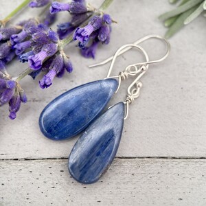 K&ouml;nnte beinhalten: Ein Paar silberne Tropfenohrringe mit blauen Kyanit-Steinen. Die Ohrringe h&auml;ngen an einer wei&szlig;en Oberfl&auml;che mit Lavendelbl&uuml;ten im Hintergrund.