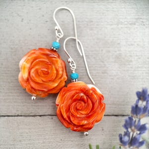 May include: A pair of orange rose-shaped earrings with silver hooks and turquoise beads. The earrings are carved with intricate details, showcasing the petals of the roses. The background is a light gray wooden surface.