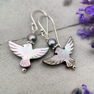 May include: A pair of silver earrings with dove-shaped pendants. The pendants have iridescent surfaces and detailed wing carvings. Each earring features a grey pearl bead and a silver hook.