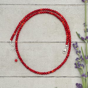 May include: A necklace made of small, round, red beads. The necklace has a silver-coloured clasp and an adjustable chain. The beads are strung together to form a double strand. The necklace is displayed on a light-coloured wooden surface.