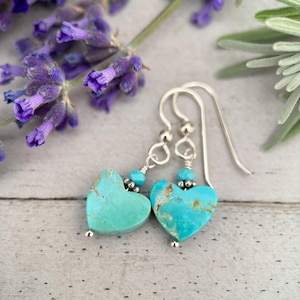 May include: Turquoise heart shaped earrings with silver hooks and beads. The earrings are on a white wooden surface with lavender flowers in the background.
