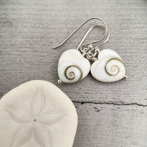 May include: A pair of heart-shaped earrings with a white base and a brown spiral design. The earrings have silver-colored hooks and small silver beads at the bottom. A white sand dollar is in the foreground.