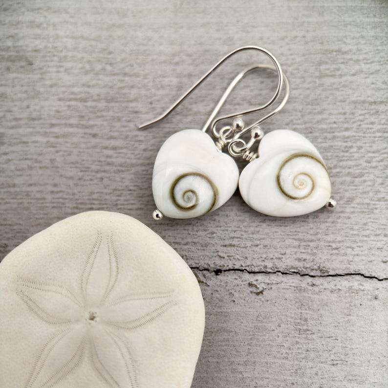 May include: A pair of heart-shaped earrings with a white base and a brown spiral design. The earrings have silver-colored hooks and small silver beads at the bottom. A white sand dollar is in the foreground.