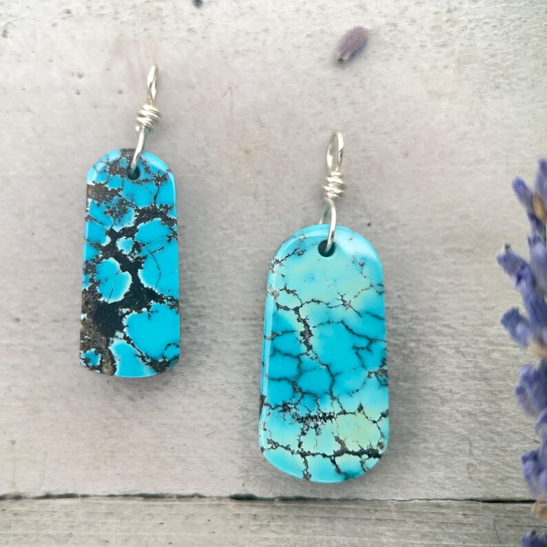 May include: Two turquoise stone pendants with silver wire loops. The rectangular pendants have a vibrant blue colour with dark brown veining. The stones are polished and have a smooth surface. The pendants are displayed on a light grey surface.