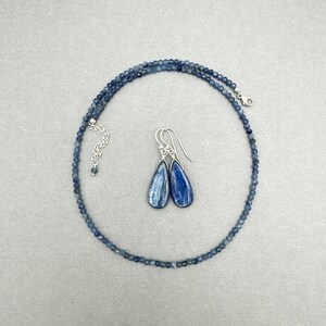 K&ouml;nnte beinhalten: Ein blaues Kyanit-Halsband und Ohrringe-Set. Die Halskette besteht aus kleinen, runden blauen Kyanit-Perlen und hat einen silbernen Verschluss. Die Ohrringe sind tropfenf&ouml;rmig und h&auml;ngen an silbernen Haken.