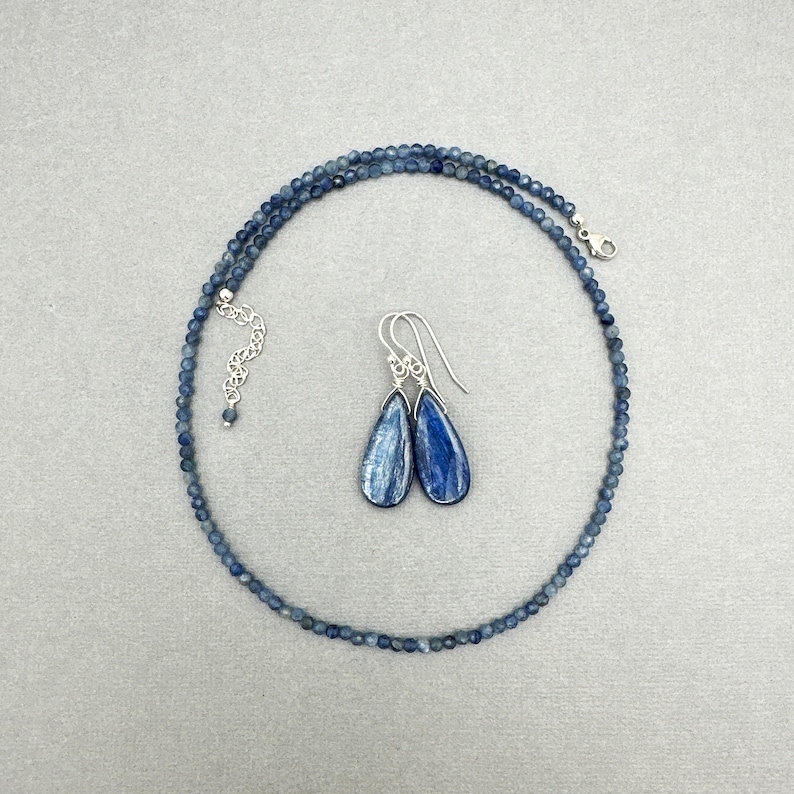 K&ouml;nnte beinhalten: Ein blaues Kyanit-Halsband und Ohrringe-Set. Die Halskette besteht aus kleinen, runden blauen Kyanit-Perlen und hat einen silbernen Verschluss. Die Ohrringe sind tropfenf&ouml;rmig und h&auml;ngen an silbernen Haken.
