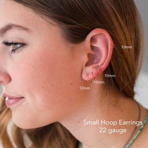 May include: A close-up of a person's ear with three gold hoop earrings. The earrings are 10mm, 10mm, and 12mm in diameter. The text "Small Hoop Earrings 22 gauge" is visible below the ear.