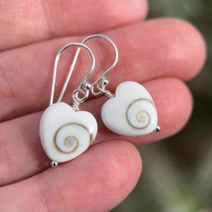 May include: A pair of heart-shaped earrings with white shells and silver hooks. Each earring features a white heart with a spiral design in shades of brown and tan. The earrings are suspended from silver hooks.