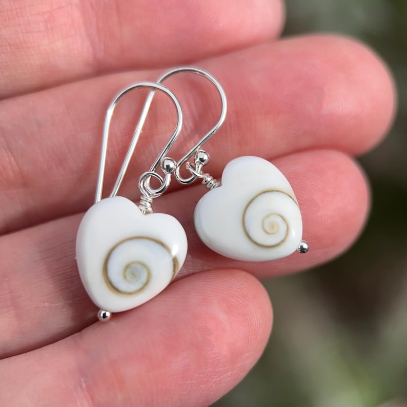 May include: A pair of heart-shaped earrings with white shells and silver hooks. Each earring features a white heart with a spiral design in shades of brown and tan. The earrings are suspended from silver hooks.