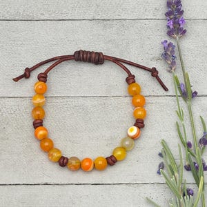 Op de afbeelding: Verstelbare armband met oranje en gele agaat kralen. De kralen zijn geregen op een bruin leren koord met een schuifknoopsluiting. De armband wordt gepresenteerd op een lichtgrijze houten ondergrond, met takjes lavendel.