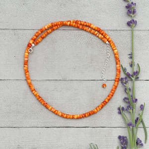 May include: An orange beaded necklace with a silver clasp and extender chain. The necklace is made of small, round beads in shades of orange and yellow. The necklace is displayed on a light gray wooden surface, with sprigs of lavender.
