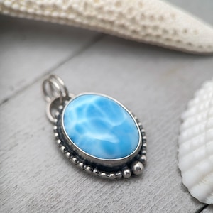 Puede incluir: Un colgante de forma ovalada que presenta una piedra azul claro lisa y pulida con patrones de veteado blanco. La piedra est&aacute; engastada en un bisel de metal plateado decorado con un borde de cuentas y una anilla doble en la parte superior.
