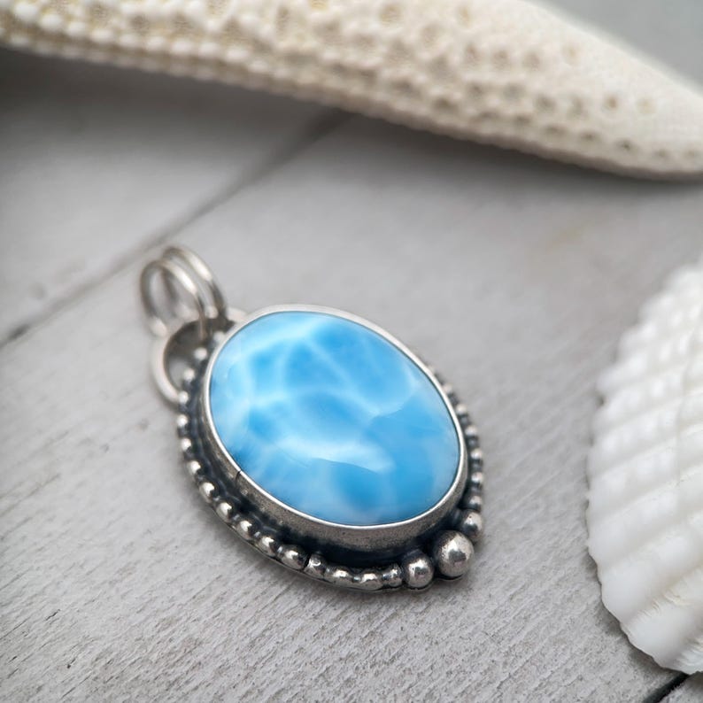 Puede incluir: Un colgante de forma ovalada que presenta una piedra azul claro lisa y pulida con patrones de veteado blanco. La piedra est&aacute; engastada en un bisel de metal plateado decorado con un borde de cuentas y una anilla doble en la parte superior.