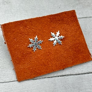 May include: A pair of silver-toned stud earrings shaped like six-pointed snowflakes with intricate, pointed edges. The earrings are displayed on a rectangular piece of textured, reddish-brown material, showcasing a metallic finish and symmetrical design.