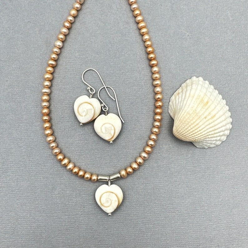 May include: A pearl necklace and matching earrings set with heart-shaped pendants. The pendants and earrings feature a white shell design with a spiral pattern. A seashell is placed to the right of the jewelry set.