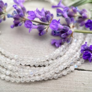 Puede incluir: Una hebra de cuentas de piedra lunar blanca con un brillo iridiscente, descansando sobre una superficie de madera con flores de lavanda.