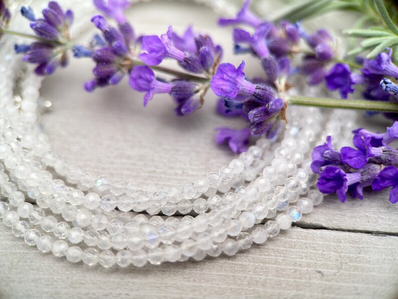 Puede incluir: Una hebra de cuentas de piedra lunar blanca con un brillo iridiscente, descansando sobre una superficie de madera con flores de lavanda.