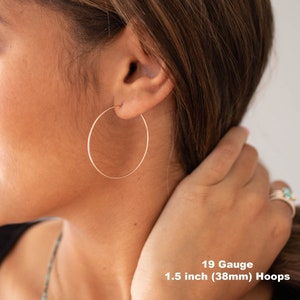 May include: Rose gold hoop earrings, 1.5 inches (38mm) in diameter, are shown in a close-up view. The earrings are 19 gauge and are worn on the ear. The image shows the earrings against a background of skin and hair.