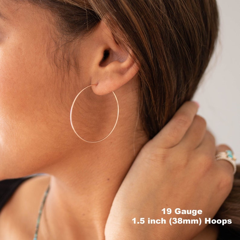 May include: Rose gold hoop earrings, 1.5 inches (38mm) in diameter, are shown in a close-up view. The earrings are 19 gauge and are worn on the ear. The image shows the earrings against a background of skin and hair.