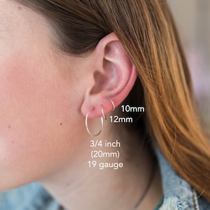 Op de afbeelding: Een oor met drie zilveren oorringen van verschillende maten. De grootste ring is 20 mm met een 19 gauge. De andere twee ringen zijn 10 mm en 12 mm. Tekst geeft de maten aan. De achtergrond is wazig.