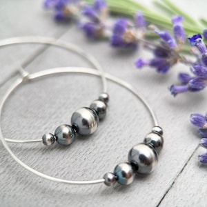 Könnte beinhalten: Silberne Creolen mit einer Reihe dunkelgrauer Perlen. Die Ohrringe sind auf einer weißen Holzoberfläche mit Lavendelblüten fotografiert.
