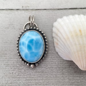 Puede incluir: Un colgante de forma ovalada con una piedra preciosa Larimar de color azul cielo pulido con patrones de veteado blanco. La piedra est&aacute; engastada en un marco de metal plateado decorado con un borde de cuentas y una anilla doble.