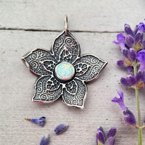 May include: A silver-colored flower pendant with an opal center stone. The pendant has detailed filigree patterns on each petal. Lavender sprigs and buds are scattered around the pendant, creating a floral theme.