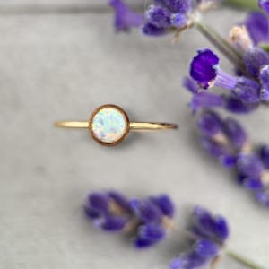 May include: A delicate gold ring with a white opal gemstone set in a wooden bezel. The ring is photographed on a white surface with lavender flowers.