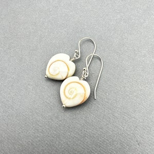 May include: A pair of heart-shaped earrings with a cream-colored, swirled design. The earrings have silver-colored hooks and small silver beads. The earrings are displayed on a gray background.