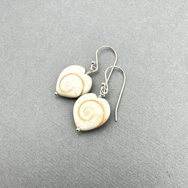 May include: A pair of heart-shaped earrings with a cream-colored, swirled design. The earrings have silver-colored hooks and small silver beads. The earrings are displayed on a gray background.