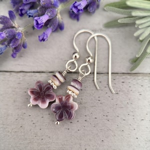 Puede incluir: Pendientes de flor de concha púrpura y blanca con ganchos de plata. Los pendientes están hechos con pequeñas cuentas de concha y una flor de concha más grande.
