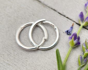 May include: A pair of small, silver hoop earrings are interlocked, set against a light grey wooden surface. Lavender sprigs with purple flowers and green stems are in the background. The earrings are a simple, elegant design, adding a touch of colour and natural beauty.