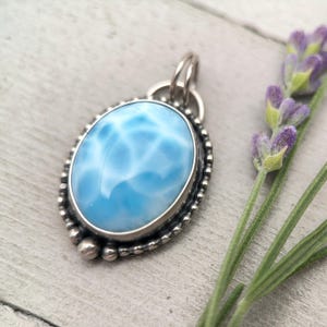 Puede incluir: Un colgante de forma ovalada con una piedra preciosa Larimar de color azul claro suave con patrones blancos similares a nubes. La piedra est&aacute; engastada en un bisel de plata pulida, rodeada por un borde de cuentas de plata y una anilla.