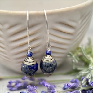May include: A pair of silver dangle earrings with blue lapis lazuli gemstone beads. The beads are round and have a silver trim. The earrings are hanging from a silver hook.