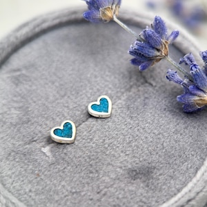 May include: A pair of heart-shaped stud earrings featuring a silver-toned metal border and a textured, bright turquoise blue inlay. The earrings are positioned on a soft, grey, circular velvet surface next to a sprig of dried purple lavender.